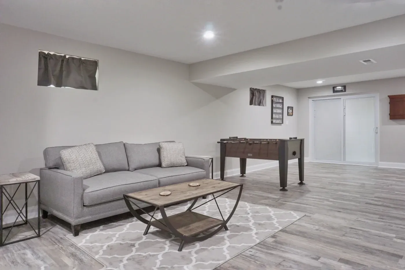 Spacious modern basement with gray sofa, patterned rug, and wooden coffee table. Foosball table and artwork on pale walls create a cozy, inviting atmosphere.