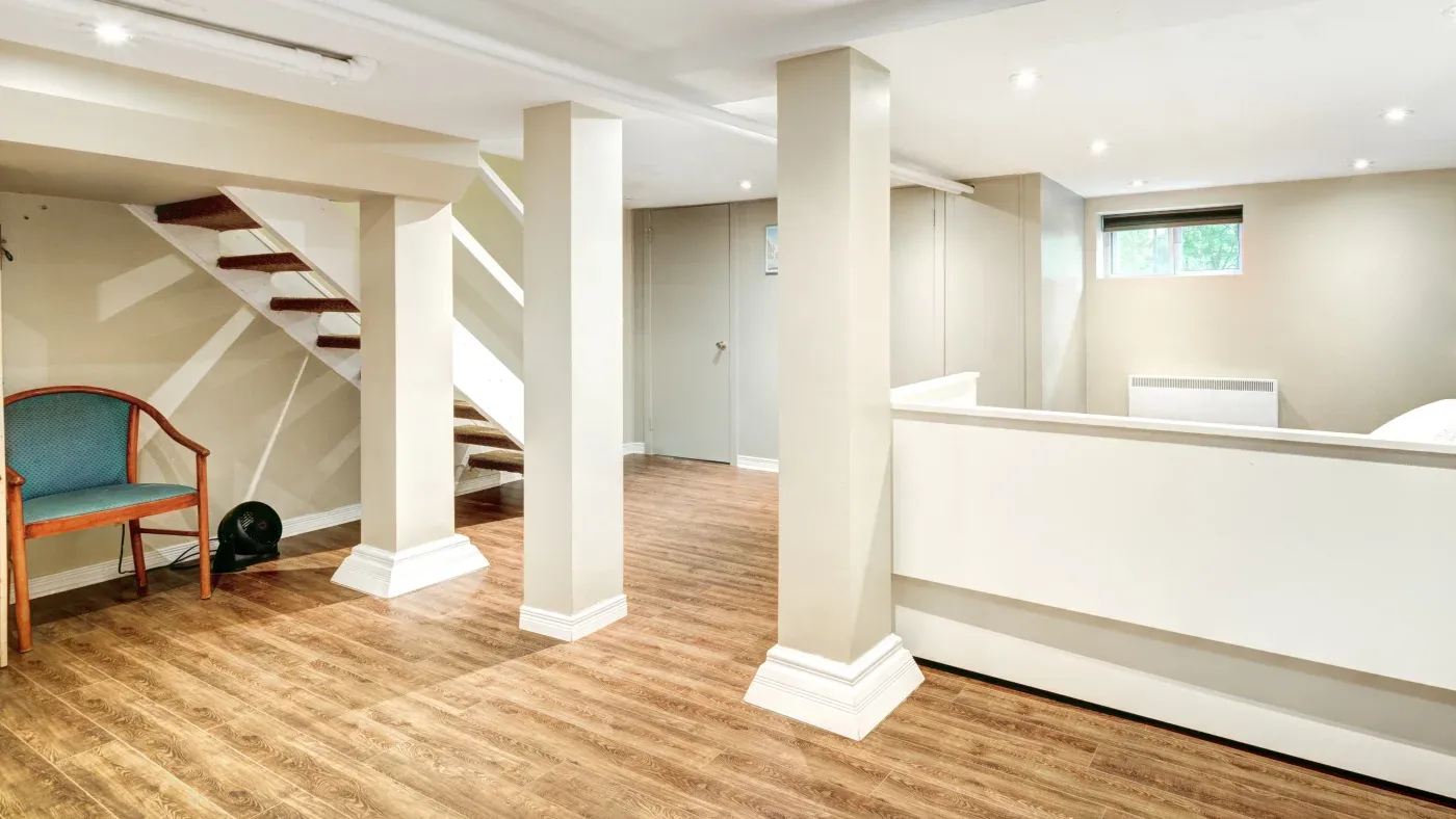 A spacious basement with wooden floors, beige walls, and white accents. A staircase, chair, and small window create a bright, inviting atmosphere.