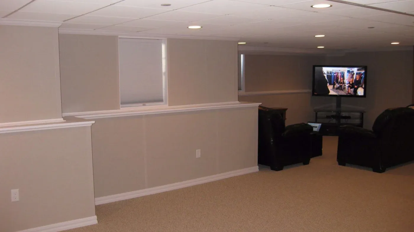 A spacious basement with beige walls and carpet, featuring a TV displaying a crowd scene, and two black leather couches creating a cozy, relaxed atmosphere.