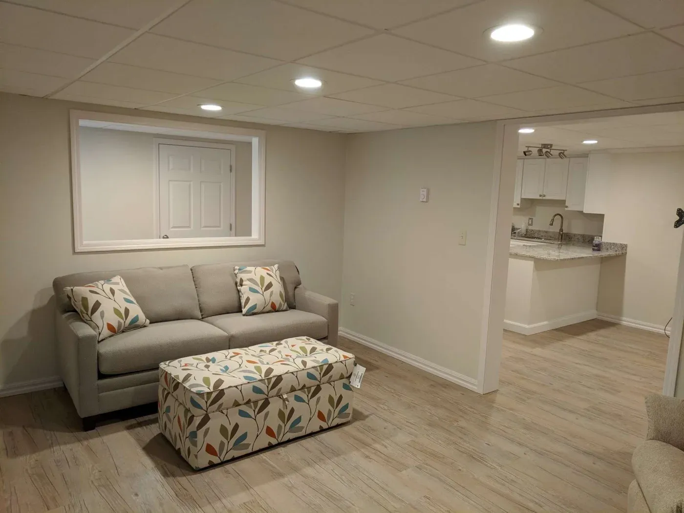 A cozy basement living room features a gray couch with floral pillows, a patterned ottoman, and light wood flooring. Bright, inviting atmosphere.