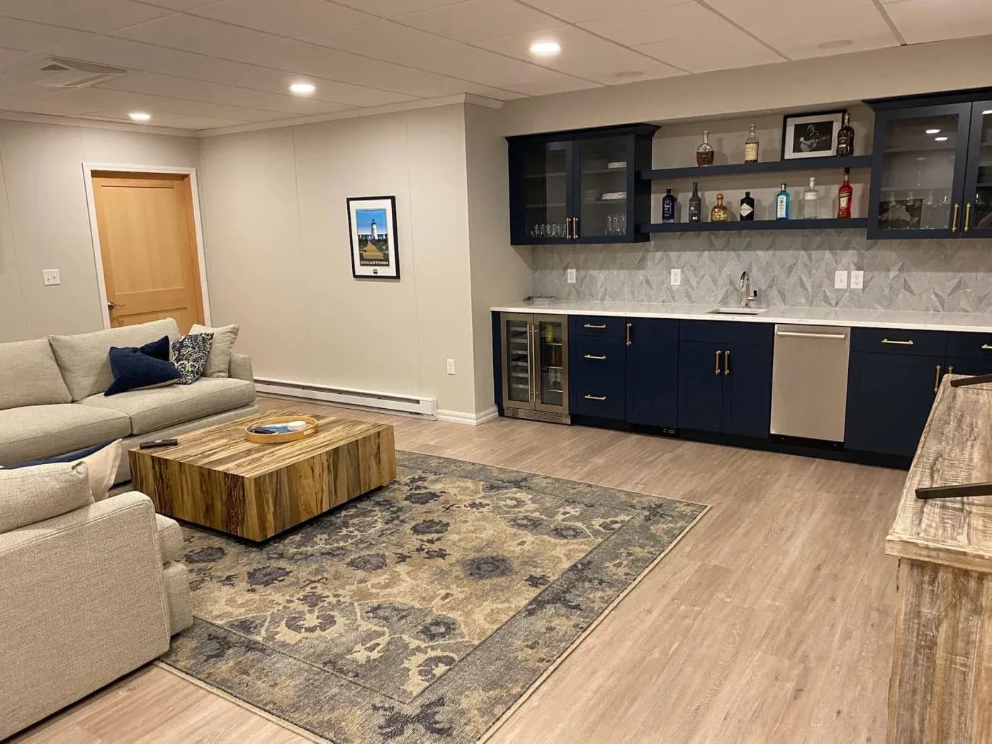 Stylish basement bar area with a modern, blue cabinet kitchenette, wooden countertops, cozy gray sofa, and patterned rug on light wood floors.