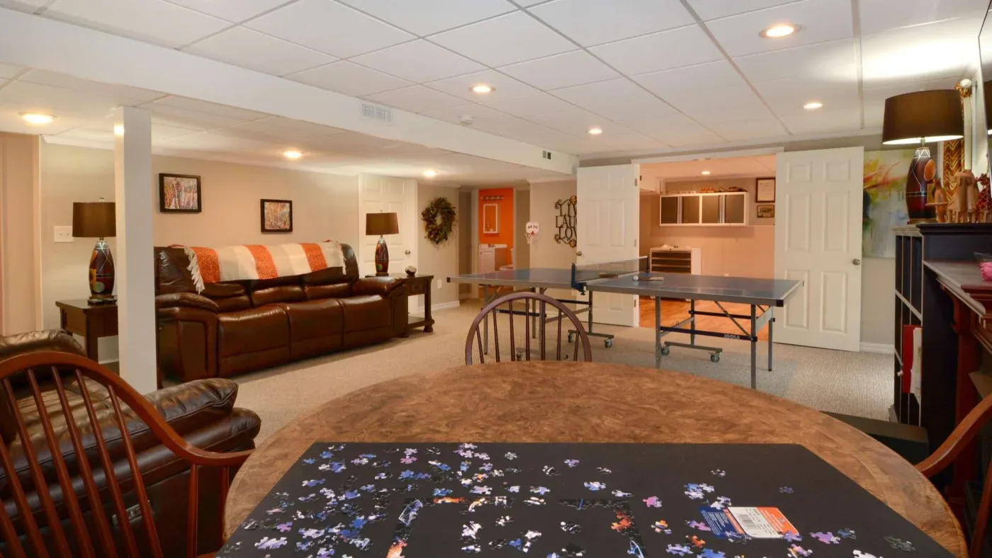 A cozy basement recreation room features a leather sofa with cushions, a table with a puzzle, a ping pong table, and warm lighting, creating a relaxed atmosphere.