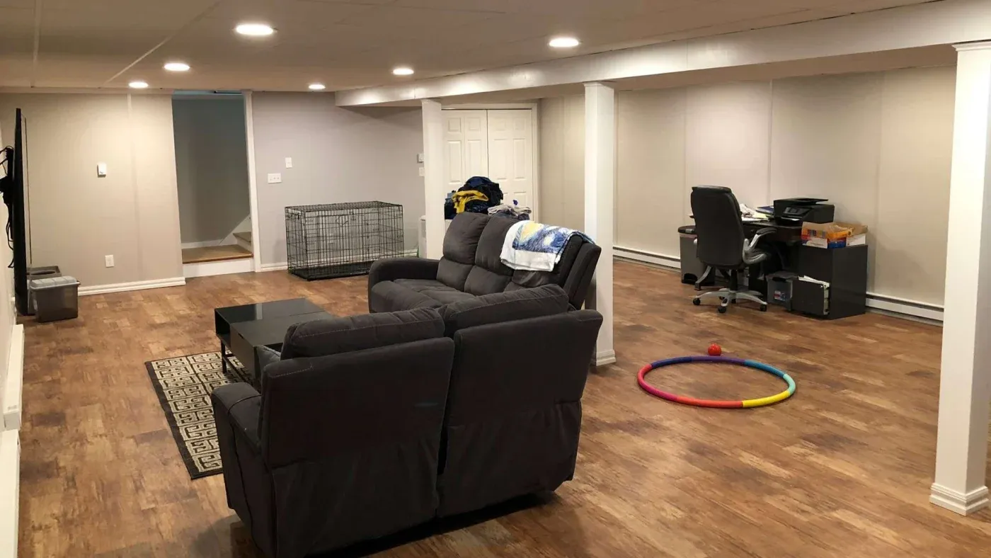 Spacious finished basement with wood flooring, featuring a dark gray sectional sofa, a TV, an office desk, and a colorful hula hoop on the floor.