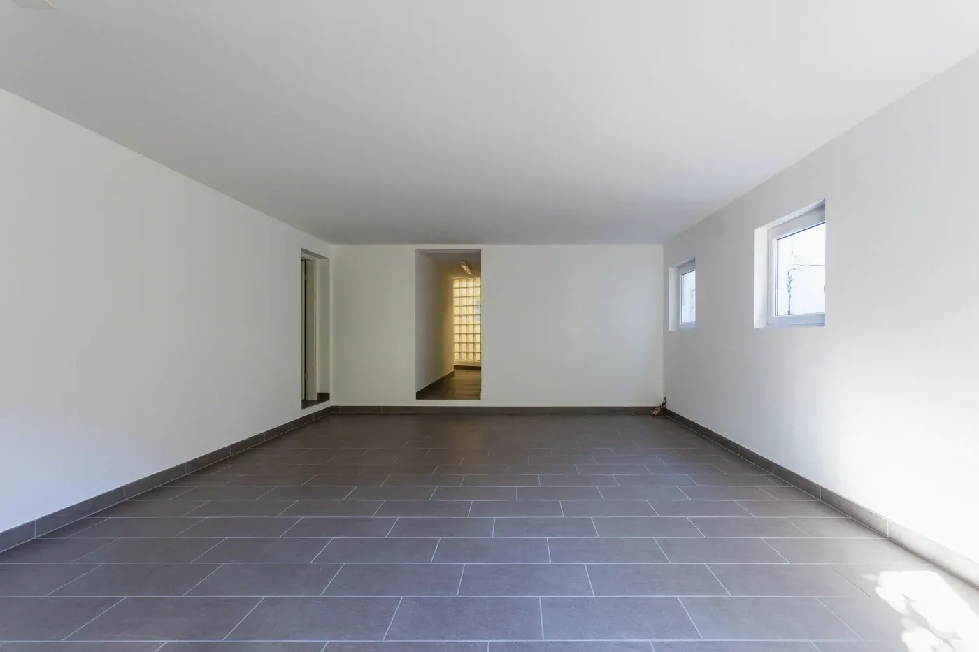 Spacious, empty room with gray tiled floor, white walls, and two small windows on the right. A doorway leads to a well-lit adjacent area. Minimalist and clean.