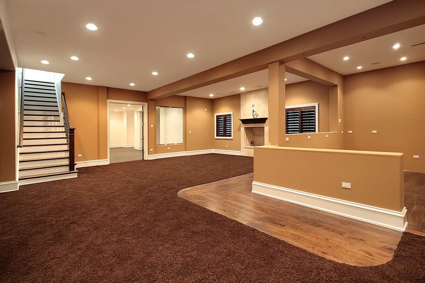 Spacious carpeted basement with warm brown walls, recessed lighting, a staircase leading up, and a fireplace, creating a cozy atmosphere.