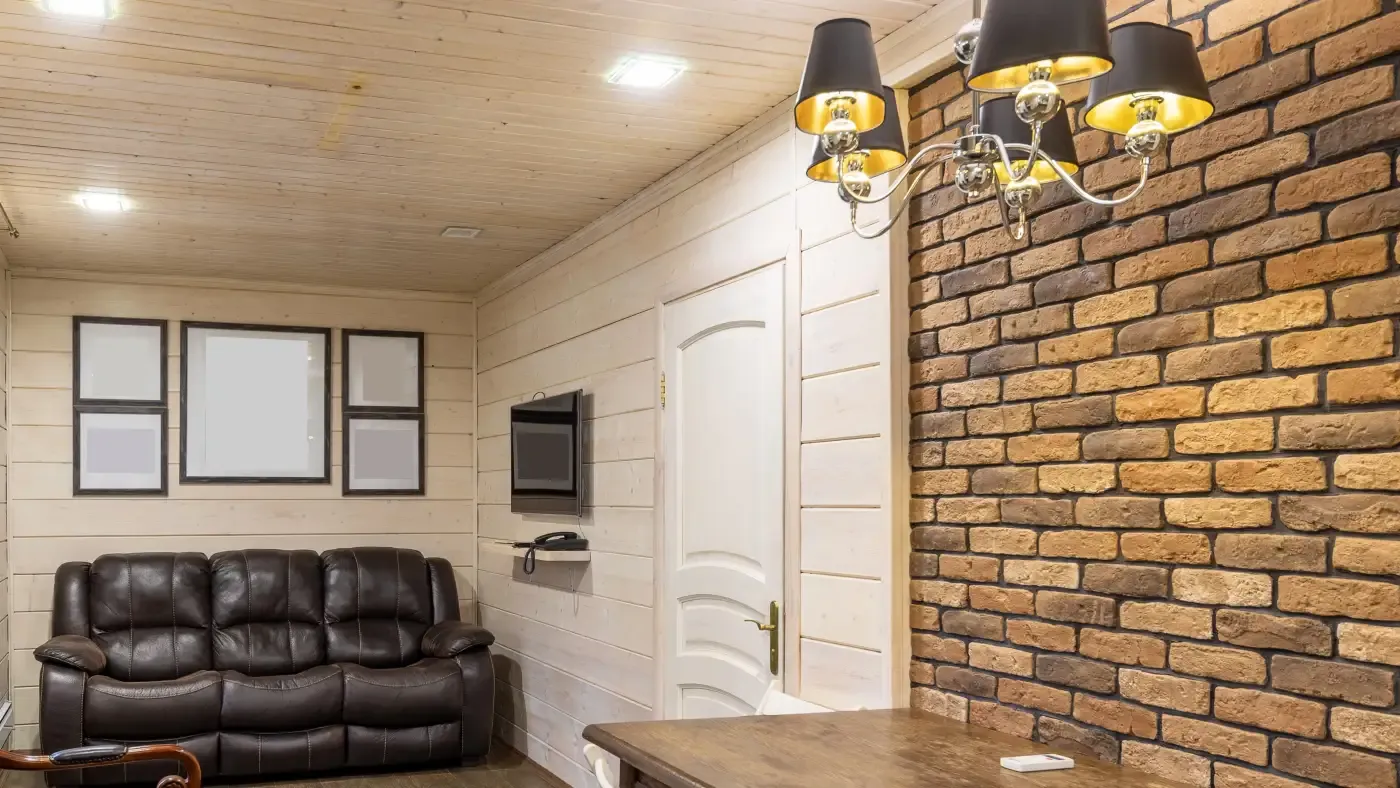 Cozy room with a rustic feel; features a brown leather sofa, brick accent wall, and warm lighting from a chandelier. A TV is mounted near a white door.
