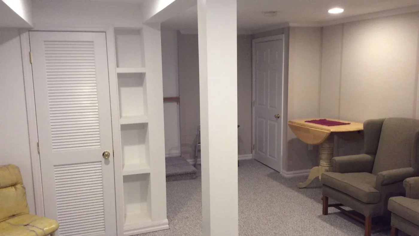 A cozy basement with white walls and carpet. There's a closet, built-in shelves, and a round wooden table. Two gray armchairs add a homely feel.