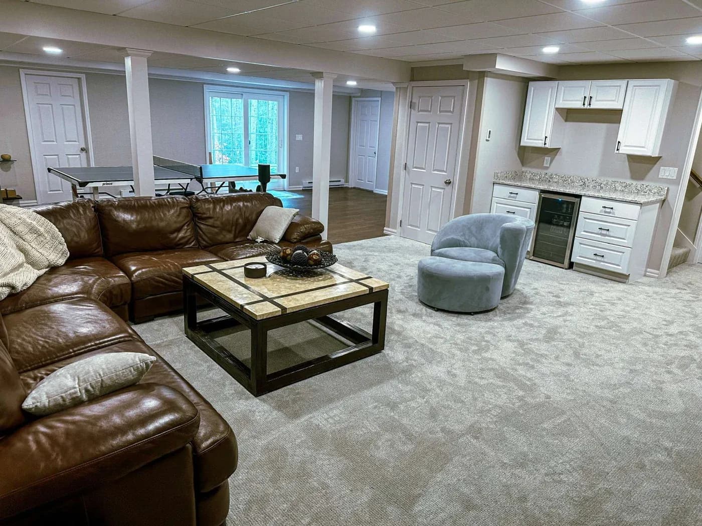 Spacious basement with a cozy brown leather sectional, a marble-topped coffee table, plush gray chair, kitchenette, and a ping pong table in the back.