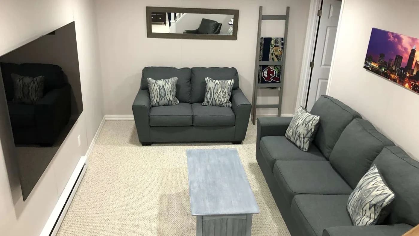A cozy living room with two gray sofas, patterned cushions, a light blue coffee table, and a flat-screen TV. A cityscape photo and a wall mirror add decor.