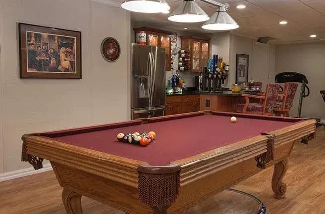 A cozy game room featuring a red pool table with balls arranged for play. Nearby, a bar area with a fridge and drinks under soft overhead lights.