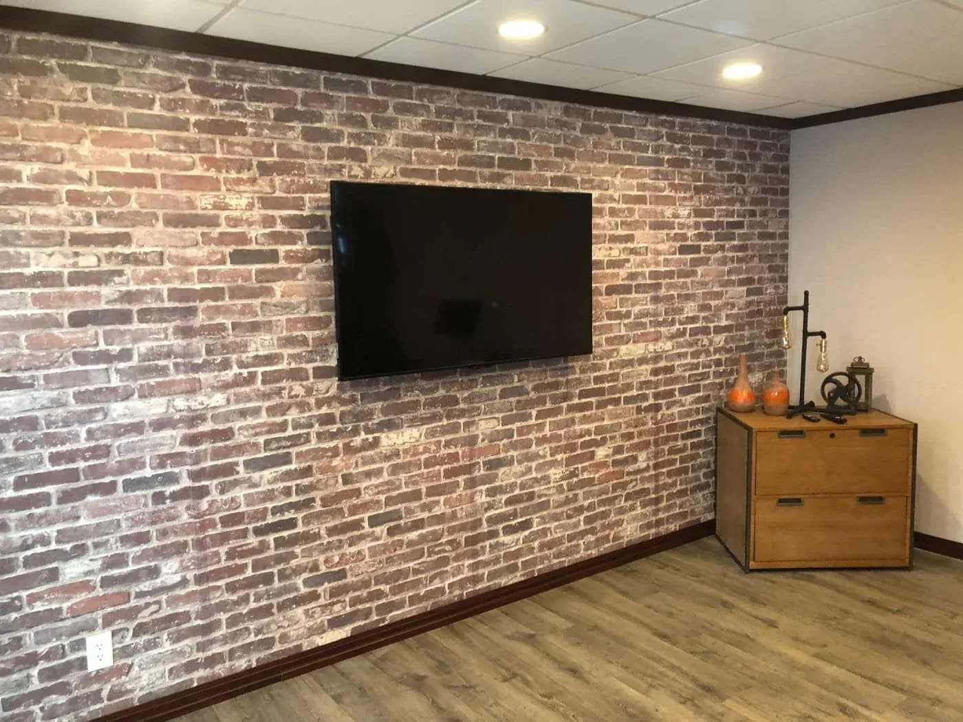 A rustic living room features a brick accent wall with a mounted flat-screen TV. A wooden cabinet holds decorative items, creating a cozy, modern feel.