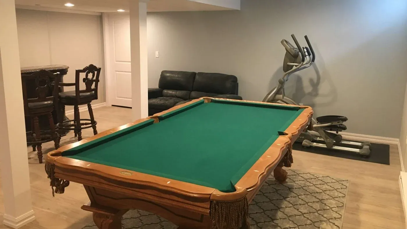 A cozy basement game room features a green-felted pool table as the centerpiece. To the right, an elliptical machine sits on a mat, and in the background, two ornate barstools stand behind a tall table. The room is softly lit with a leather sofa against a light gray wall, creating a relaxed, inviting atmosphere.
