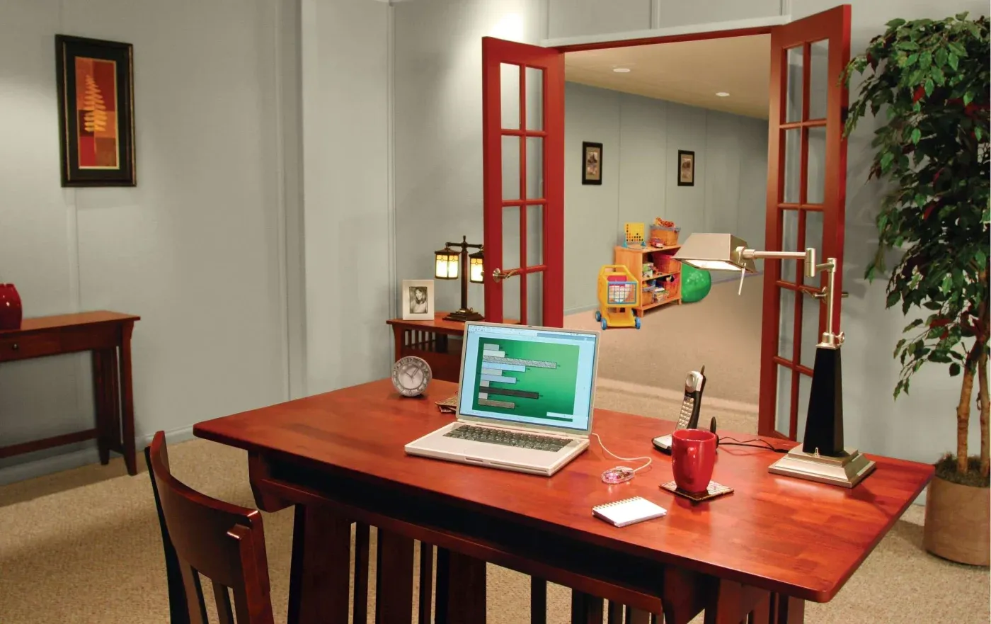 A home office with a wooden desk features a laptop displaying code, a red mug, a notebook, and a pen. The open door reveals a colorful playroom.