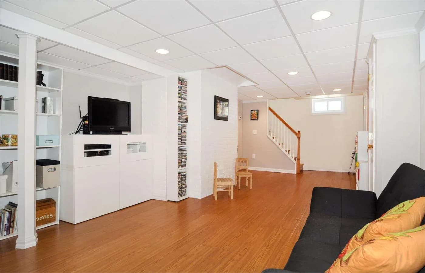 A spacious basement with wood flooring, white walls, and a ceiling with recessed lights. Features a TV, a black couch, a staircase, and shelves with boxes.