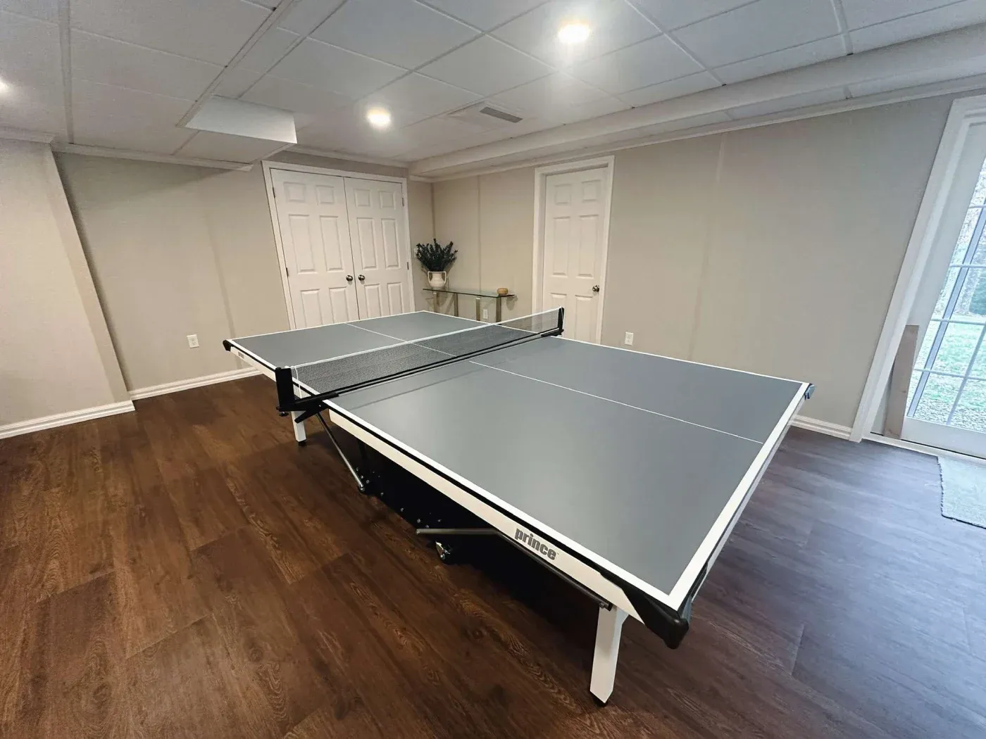 A modern basement room with a table tennis setup on a dark wood floor. The room has white walls, double doors, and large windows, creating a spacious, recreational vibe.