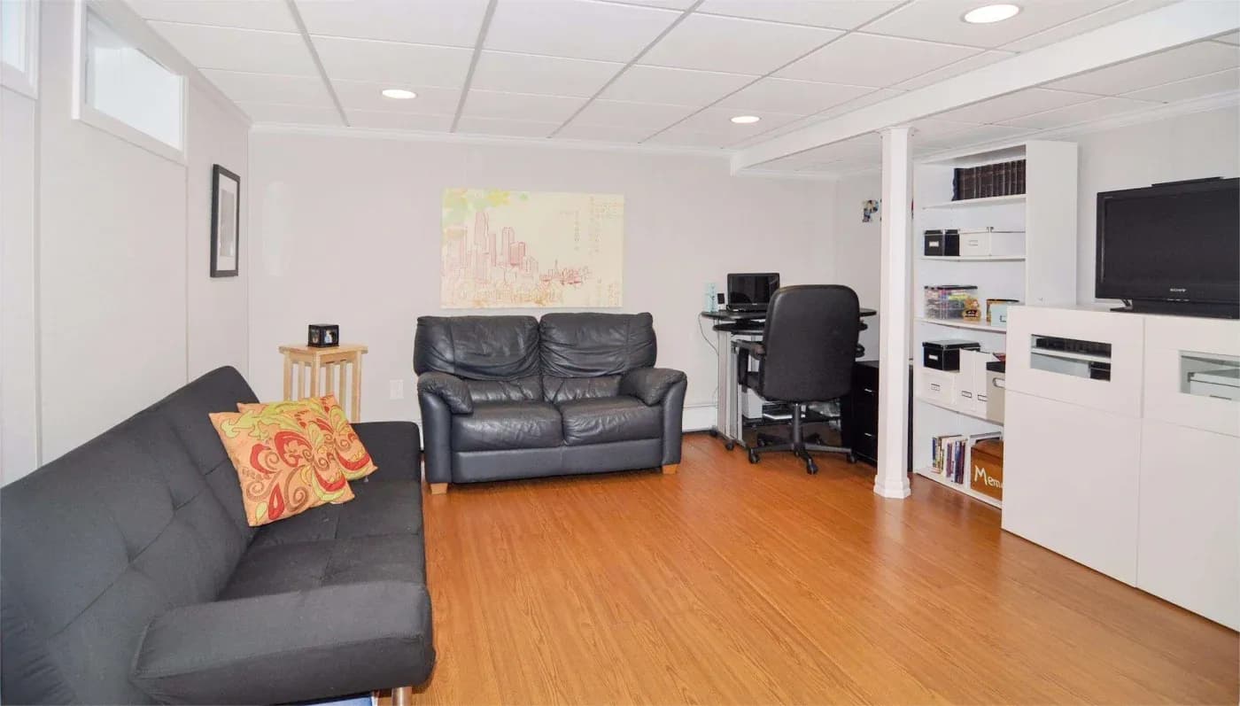 Bright, tidy basement lounge with black sofas, colorful patterned pillows, wood flooring, office desk, shelving, and wall-mounted TV.