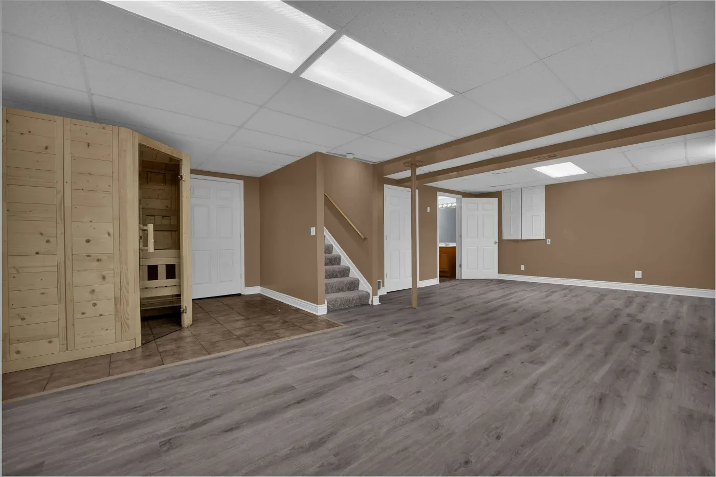 Spacious basement with gray laminate flooring and brown walls. Features a wooden sauna, carpeted stairs, and bright ceiling lights, conveying a modern, cozy vibe.
