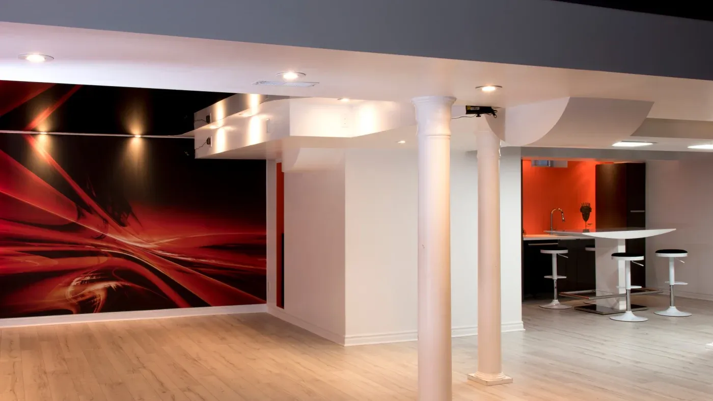 Modern basement with light wood floors, white pillars, and a striking red and black abstract wall. A sleek bar area with stools is visible, creating a stylish, contemporary ambiance.
