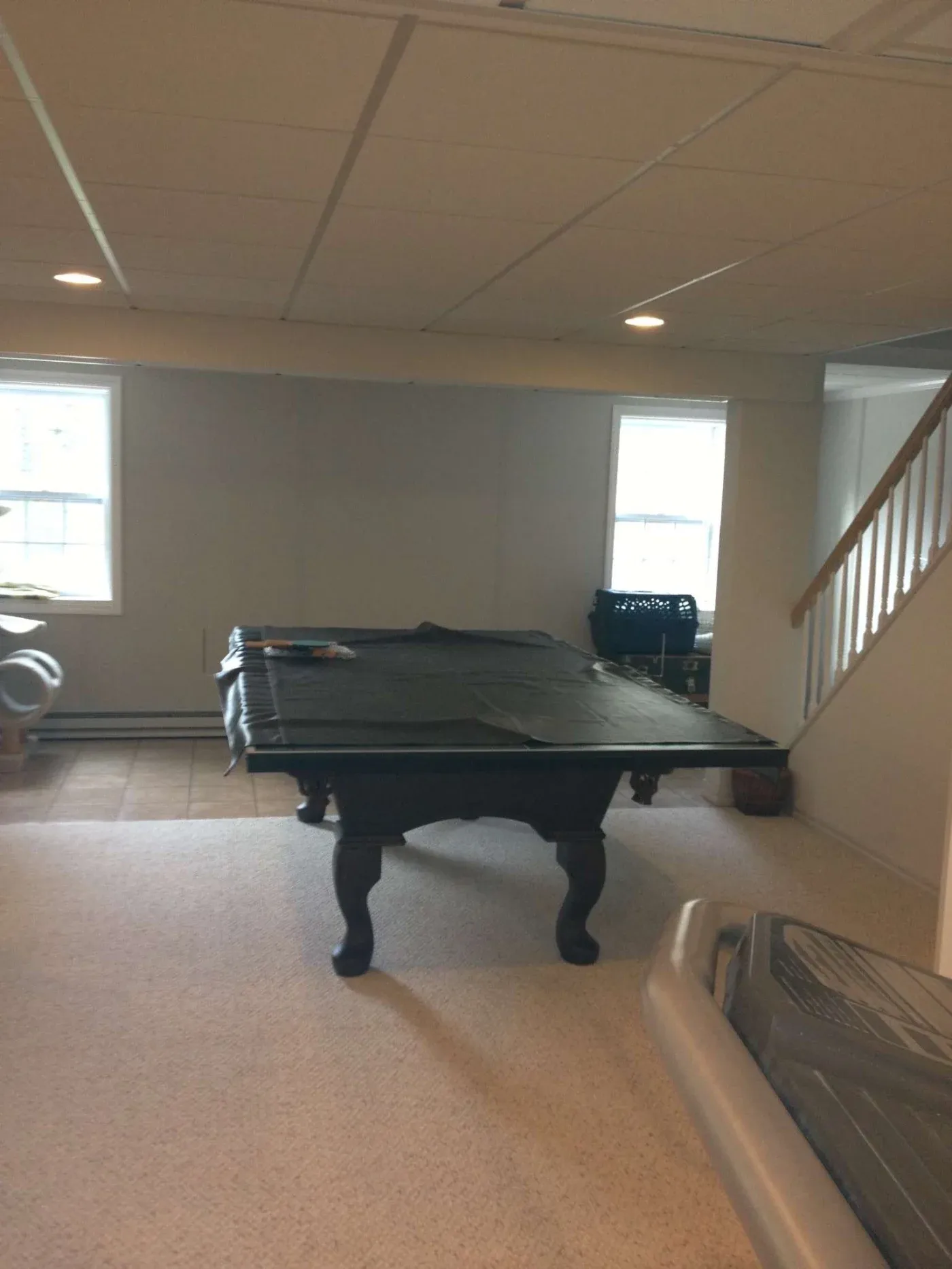 Dimly lit basement room with a pool table covered in a black cloth. Beige carpet, tiled floor near windows, and stairs lead upstairs. Cozy and quiet ambiance.