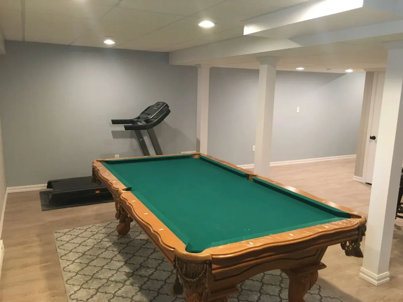 Basement recreation room with a green-felt pool table in the center. A treadmill is against the gray wall. The space is well-lit and inviting.