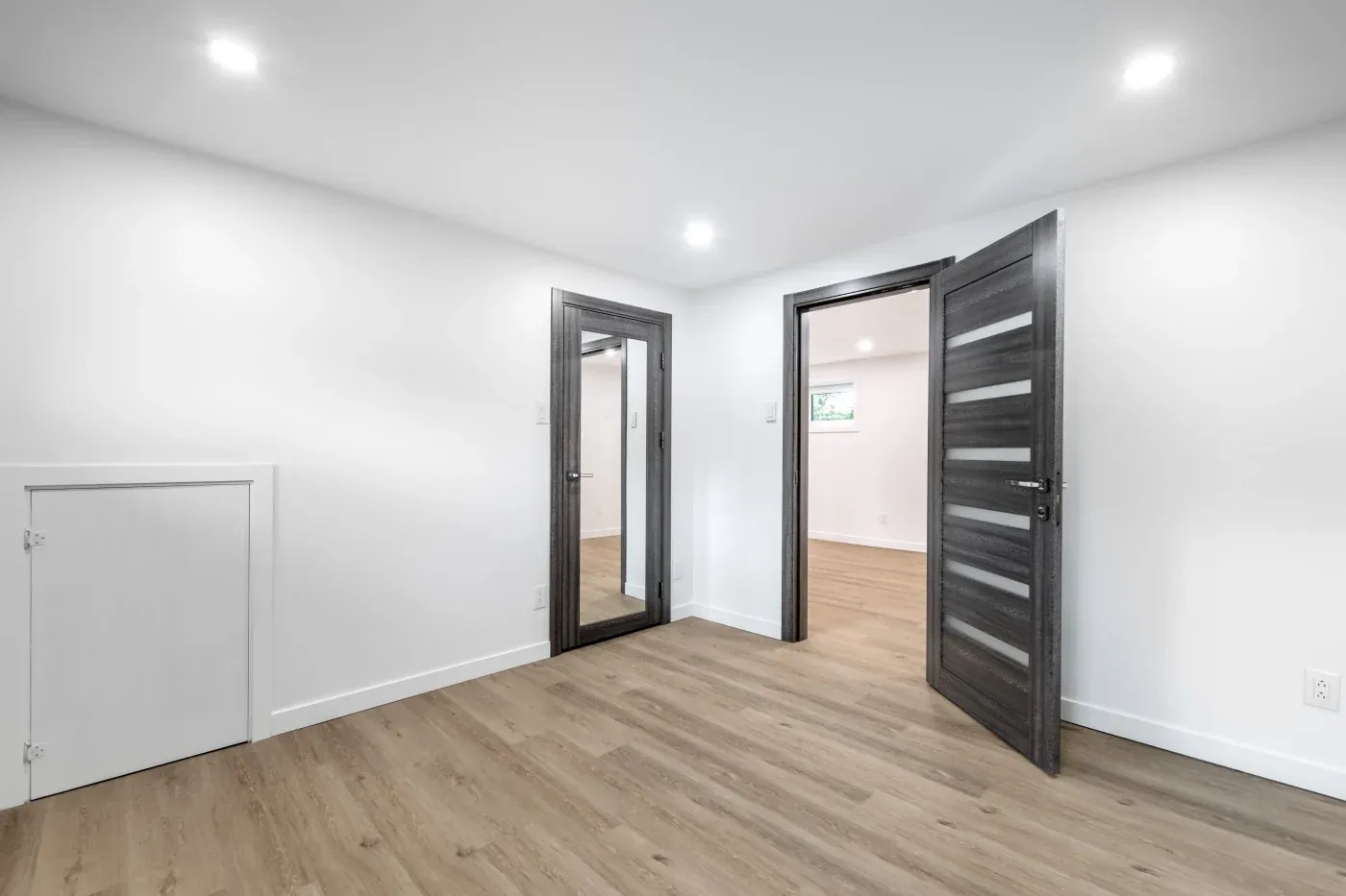 Modern room with light wooden flooring, white walls, and recessed lighting. A dark-framed door is open, revealing a bright adjacent room.