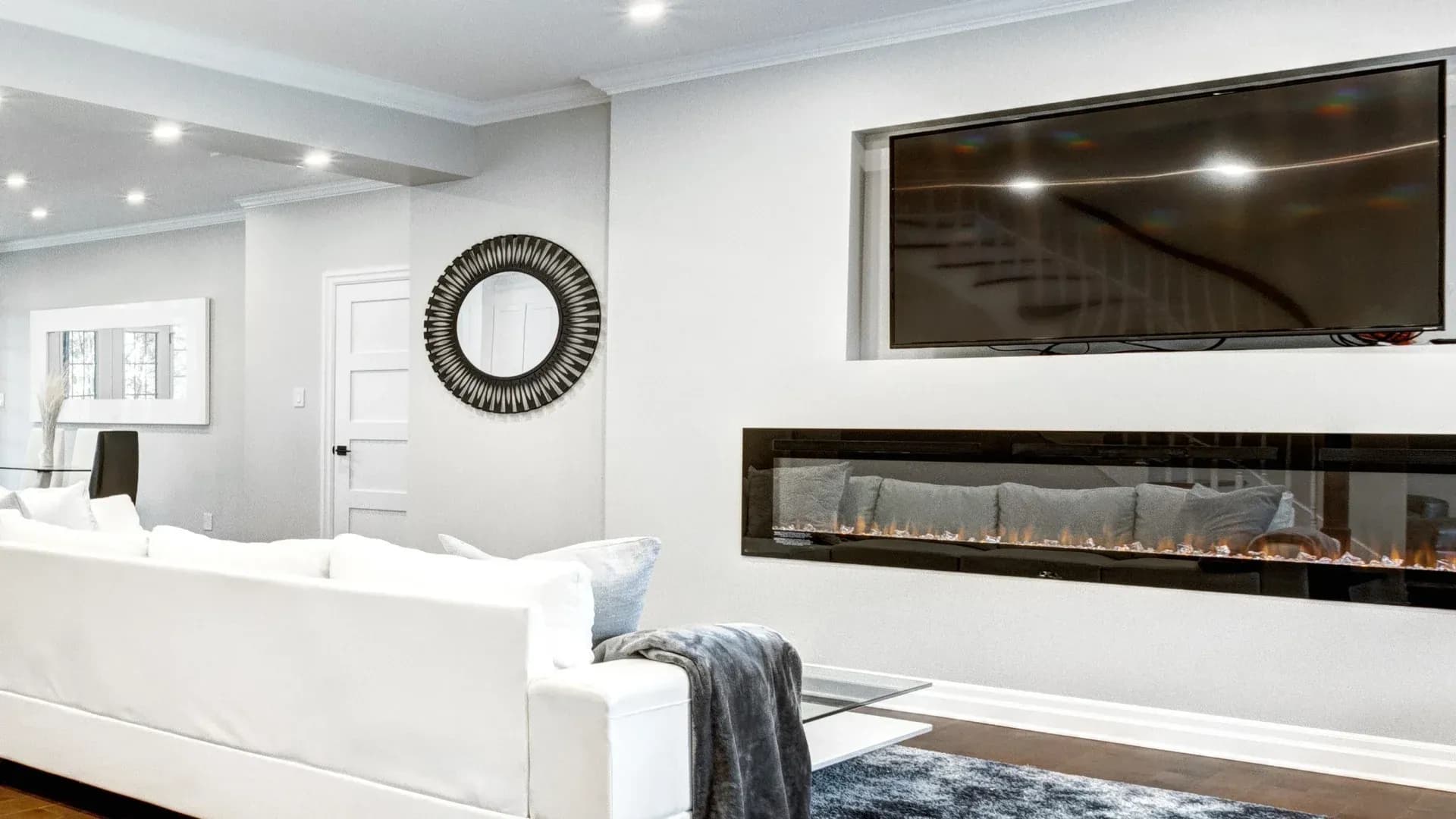 Modern living room with a sleek white sofa and a cozy gray throw. A large TV is mounted above a linear gas fireplace, radiating warmth and comfort.