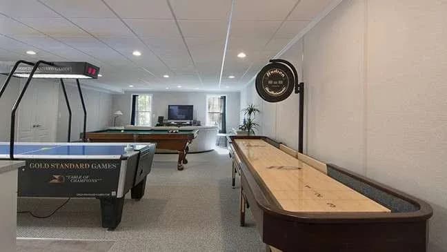 Spacious game room with a shuffleboard table, air hockey, and pool table. Bright lighting and a cozy ambiance with a TV and seating in the background.