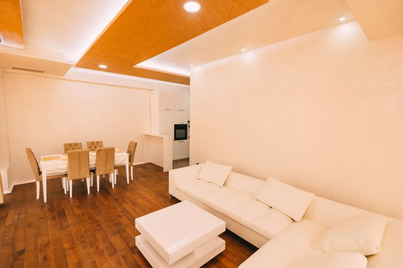 Modern living room with a white L-shaped sofa, matching coffee table, and adjacent dining area. Warm lighting and wooden floors add coziness.