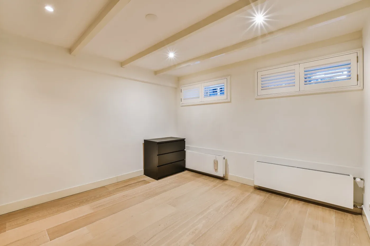 Simple, empty room with light wood flooring, white walls, and two high windows. A black dresser and radiator are along one wall. Bright lighting creates a clean, minimalist feel.