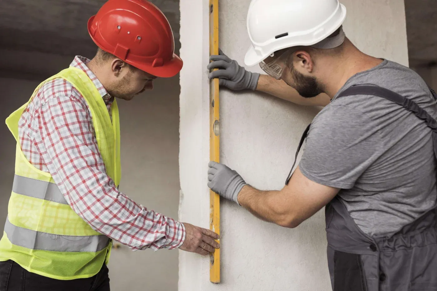 Two construction workers using a spirit level to align a wall. One wears a red helmet and safety vest, the other a white helmet and gray overalls.