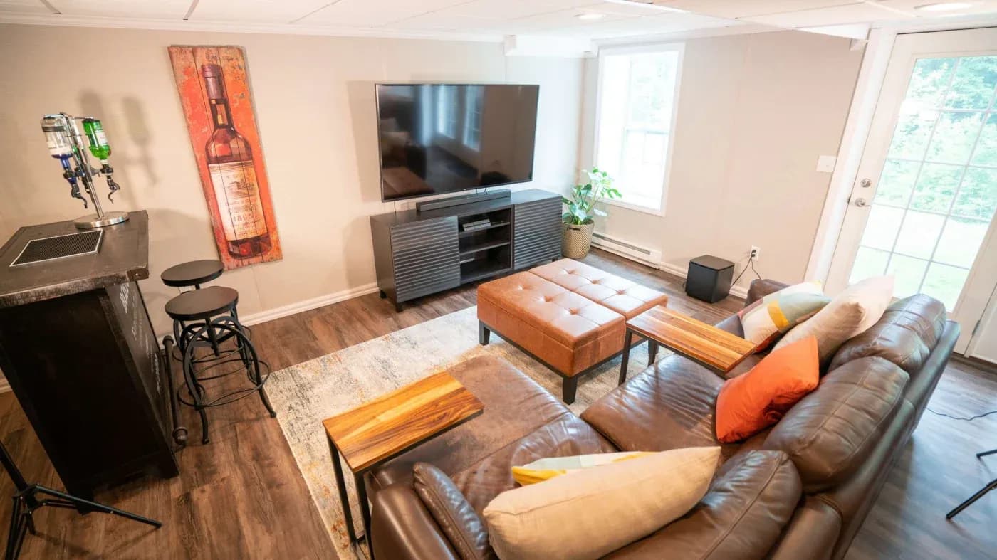 Cozy living room with a brown leather sectional, colorful cushions, wall-mounted TV, and bar area. Large windows brighten the space.