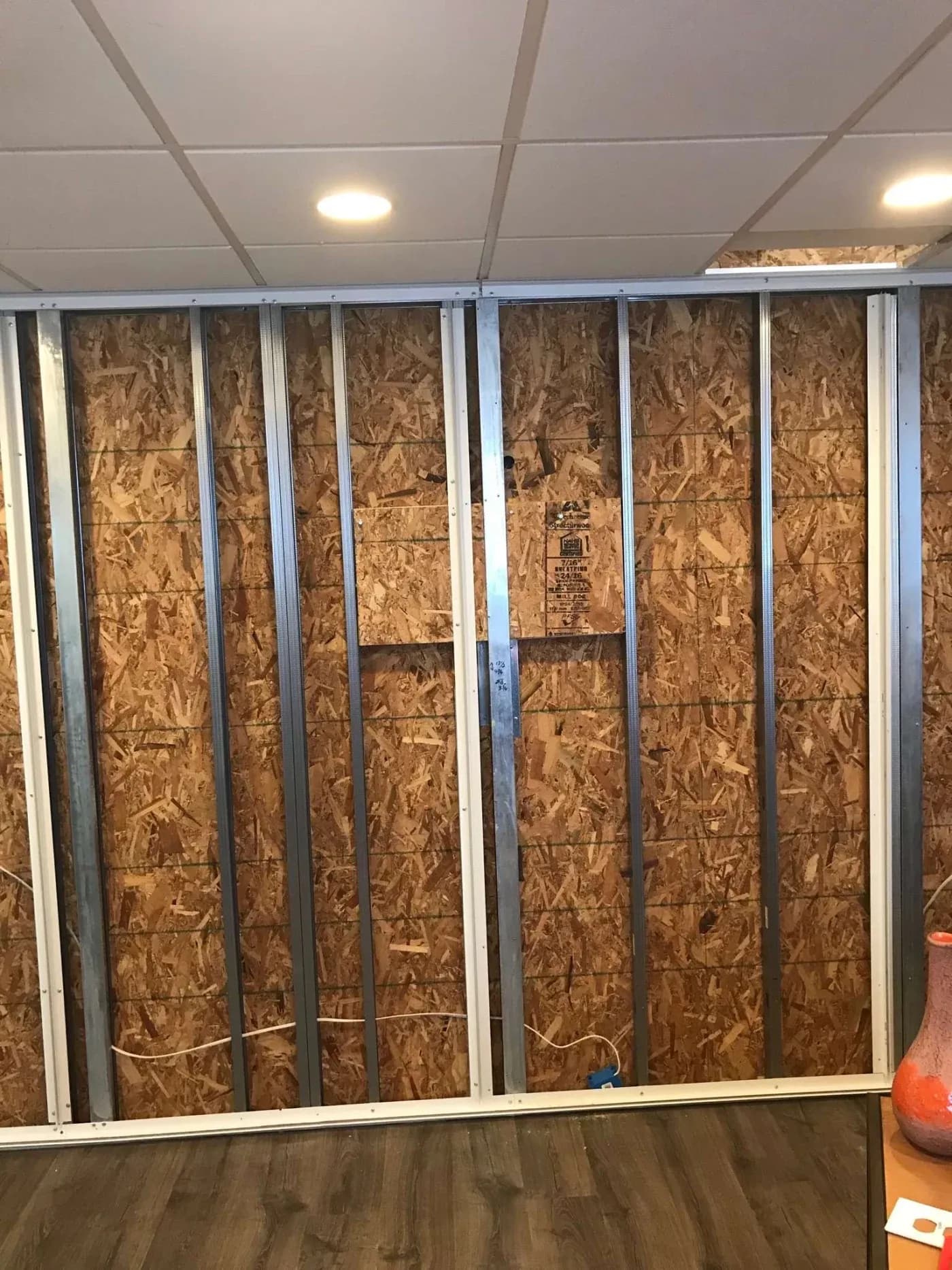 Exposed section of an unfinished wall with wooden panels and metal studs. Overhead lights illuminate the space, with a wooden floor below.