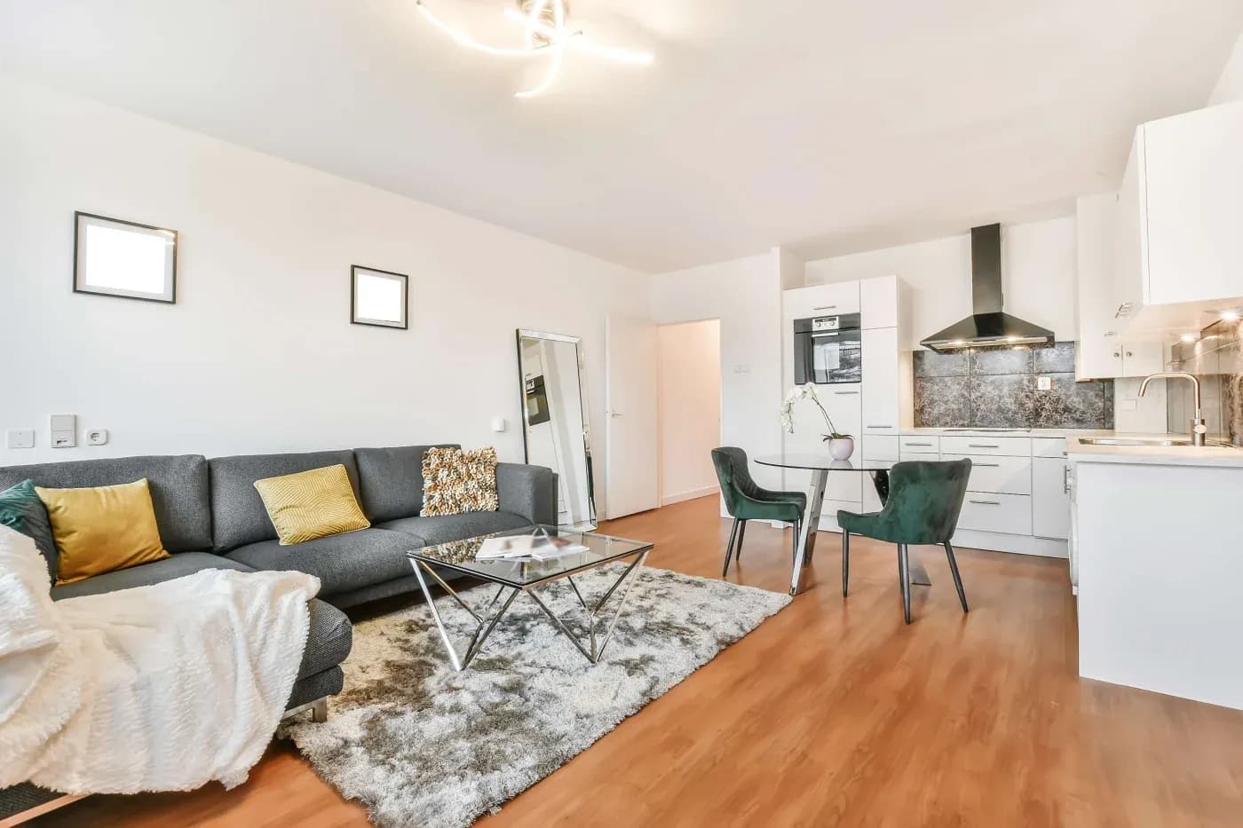 Modern open-plan living room and kitchen with a grey sectional sofa, yellow cushions, and a soft rug. Green chairs and sleek kitchen enhance a cozy, elegant vibe.