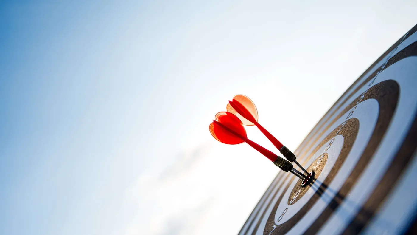 Two red darts perfectly hit the bullseye of a black and white target against a clear blue sky, symbolizing precision and success.