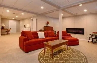 Spacious basement with red sectional sofa, coffee table, and wall-mounted fireplace. Soft lighting and neutral walls create a cozy, inviting atmosphere.
