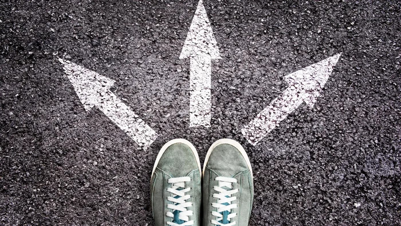 Green sneakers stand on an asphalt road facing three white arrows pointing in different directions, symbolizing choice and decision-making.