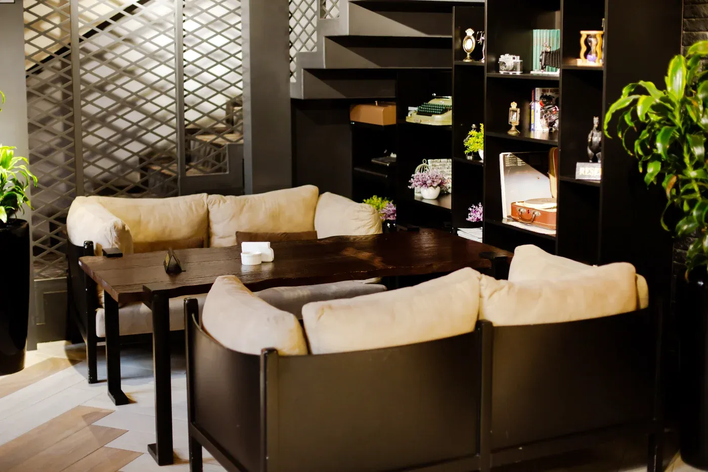 Cozy seating area with beige cushioned chairs around a dark wooden table, flanked by grids on one side and a bookshelf with plants and decor on the other.