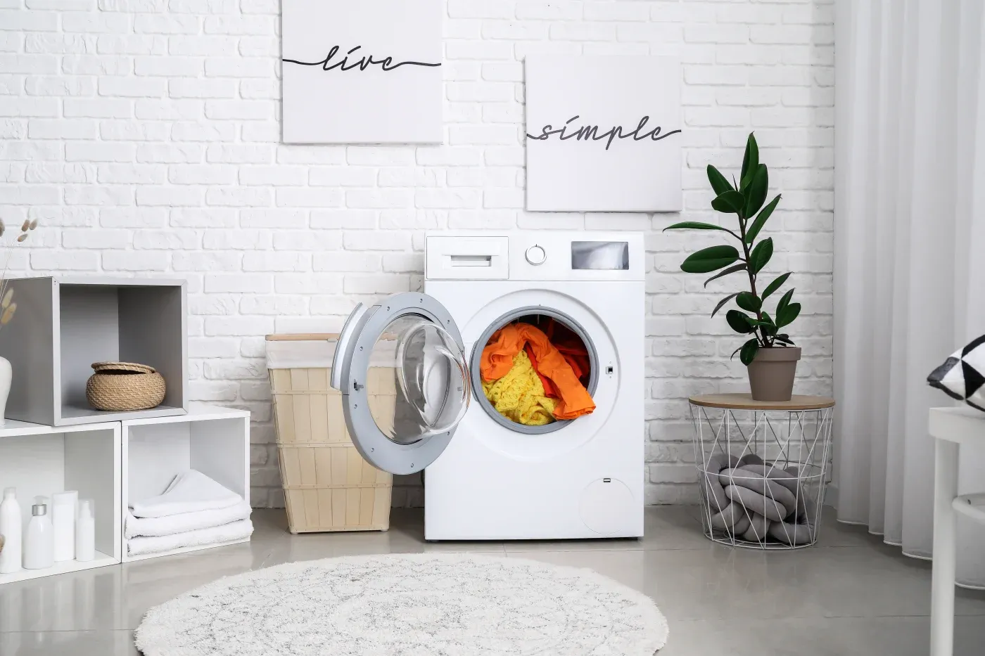 Laundry room with a modern white washing machine, open door revealing colorful clothes. Nearby are plants, baskets, and minimalist decor on white brick walls.