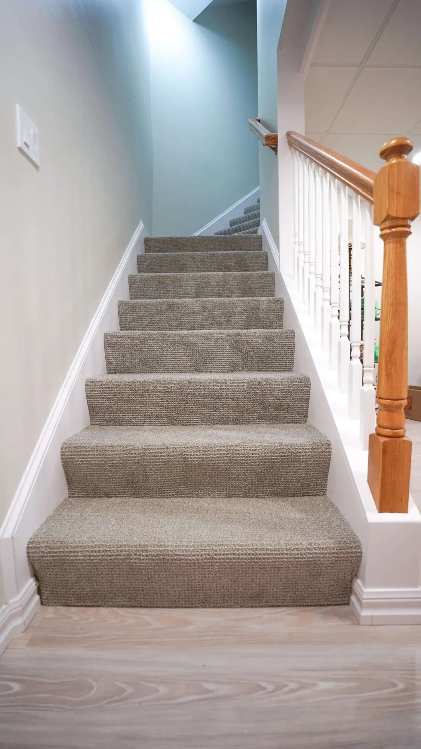 A carpeted staircase with beige steps is flanked by a wooden handrail and white balusters, leading upwards in a well-lit interior home setting.