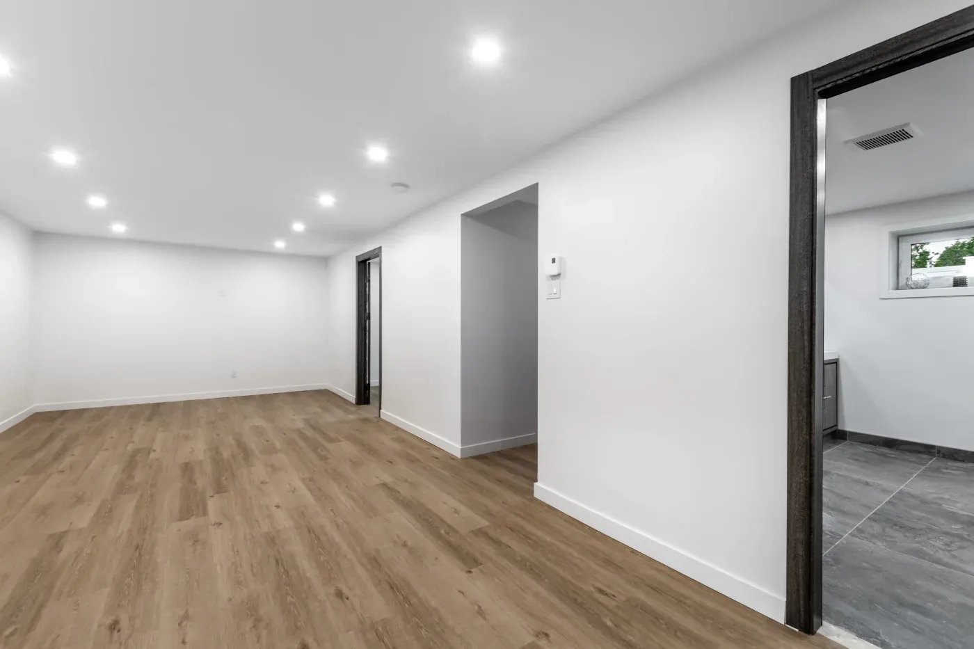 Modern basement with white walls, wood-like flooring, and recessed lighting. An open doorway leads to another room, creating a clean, spacious feel.