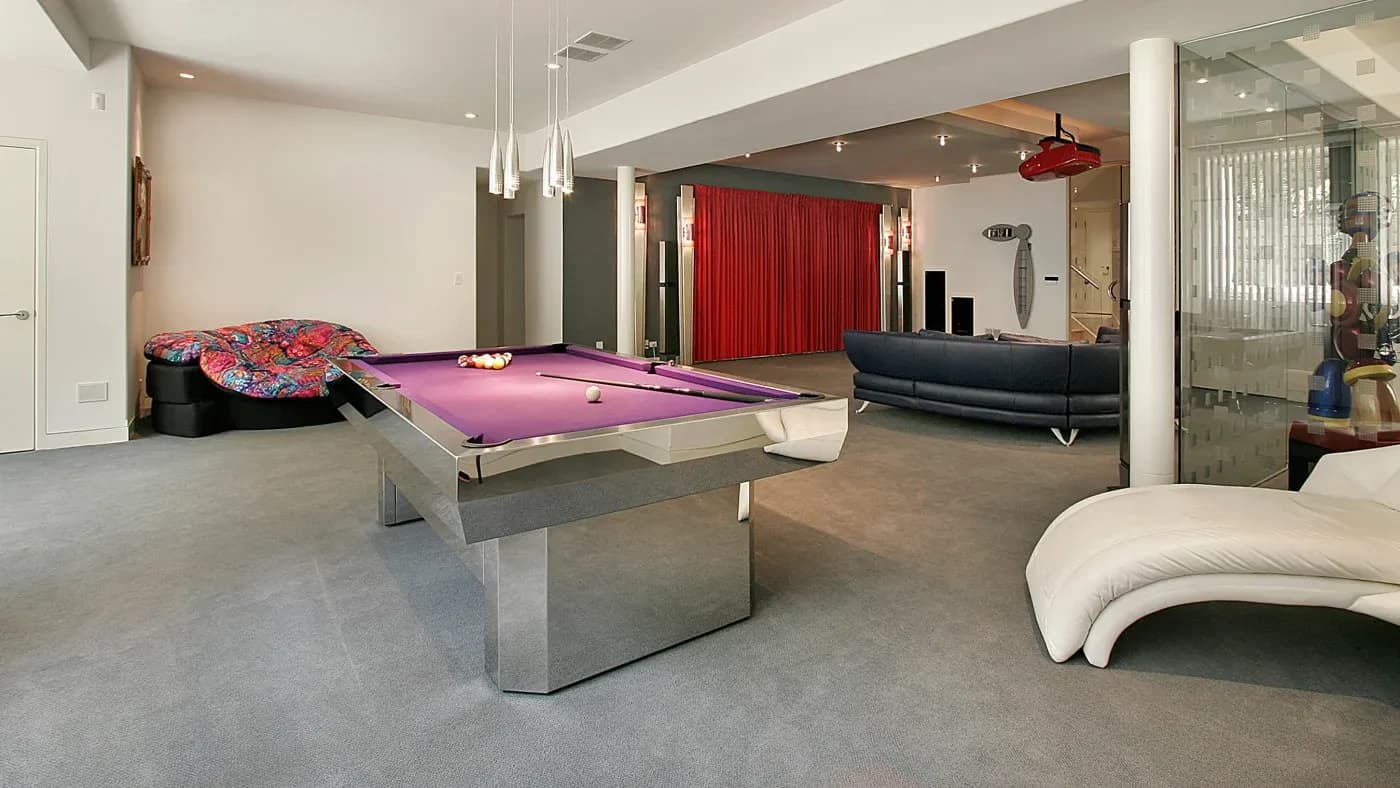 Spacious game room with a modern purple pool table, abstract art on the wall, a cozy lounge area with curved couches, and vibrant red curtains.