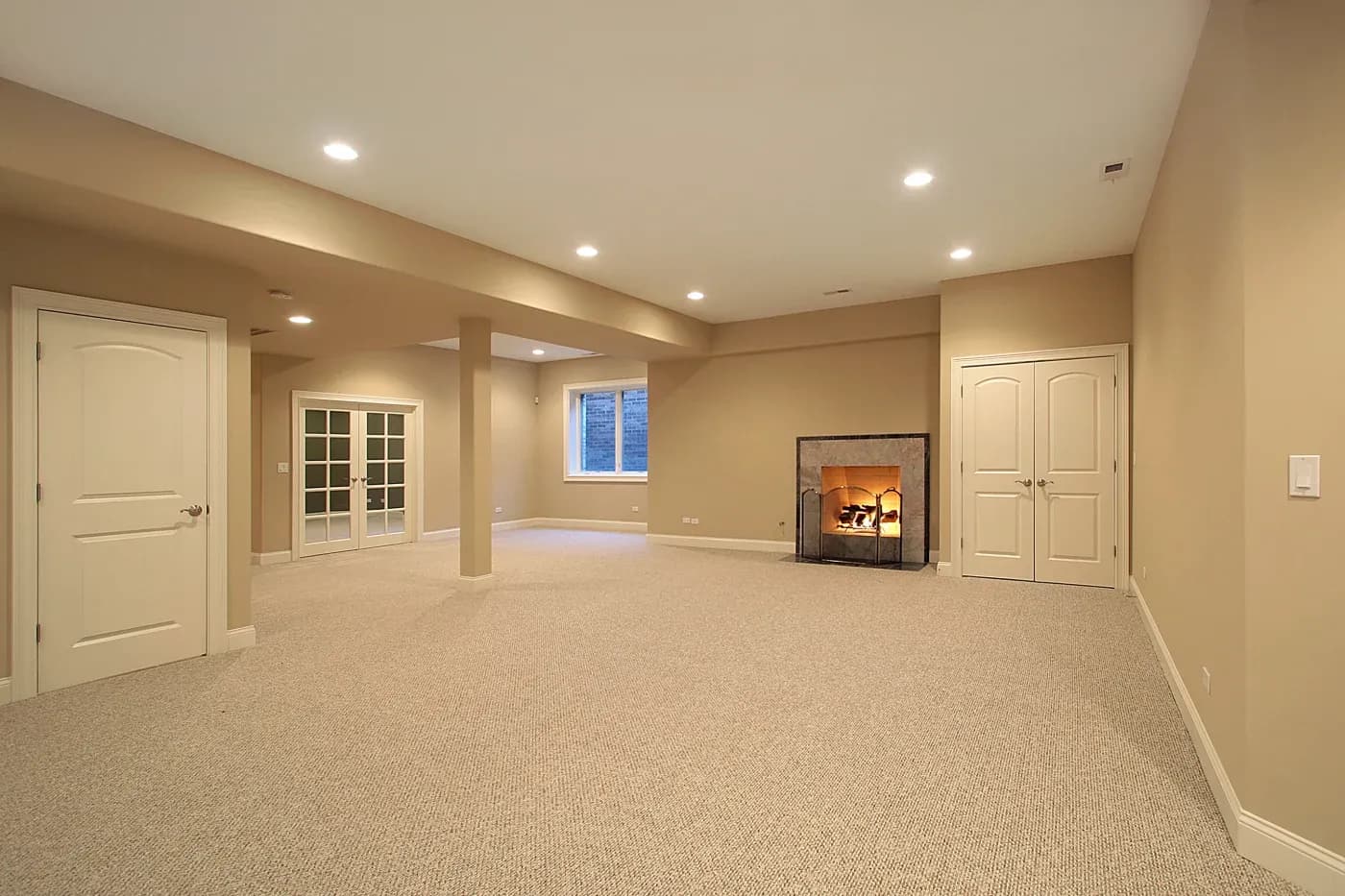 Spacious beige room with soft lighting, featuring a lit fireplace, carpet flooring, double doors, and a window. Cozy and warm ambiance.