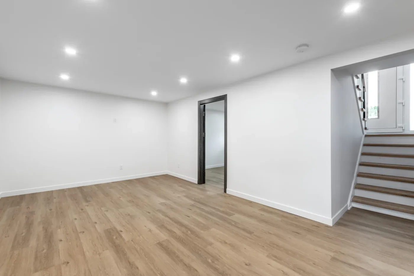 Spacious, empty room with light wood flooring and white walls. Recessed ceiling lights and a staircase on the right create a bright, open atmosphere.