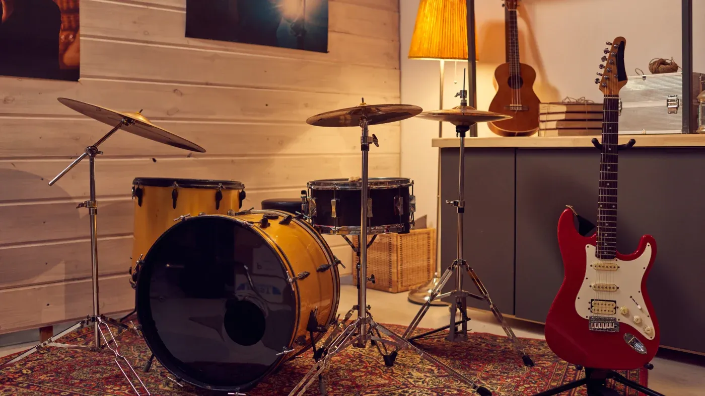 A cozy music room features a yellow drum set on a patterned rug, a red electric guitar standing nearby, and a warm-lit lamp. A ukulele is hung on the wall.
