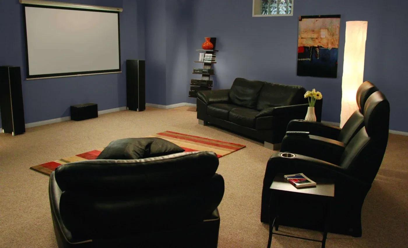 A cozy home theater with black leather seating facing a large projector screen on a blue wall. Soft lighting and a rug add warmth and comfort.