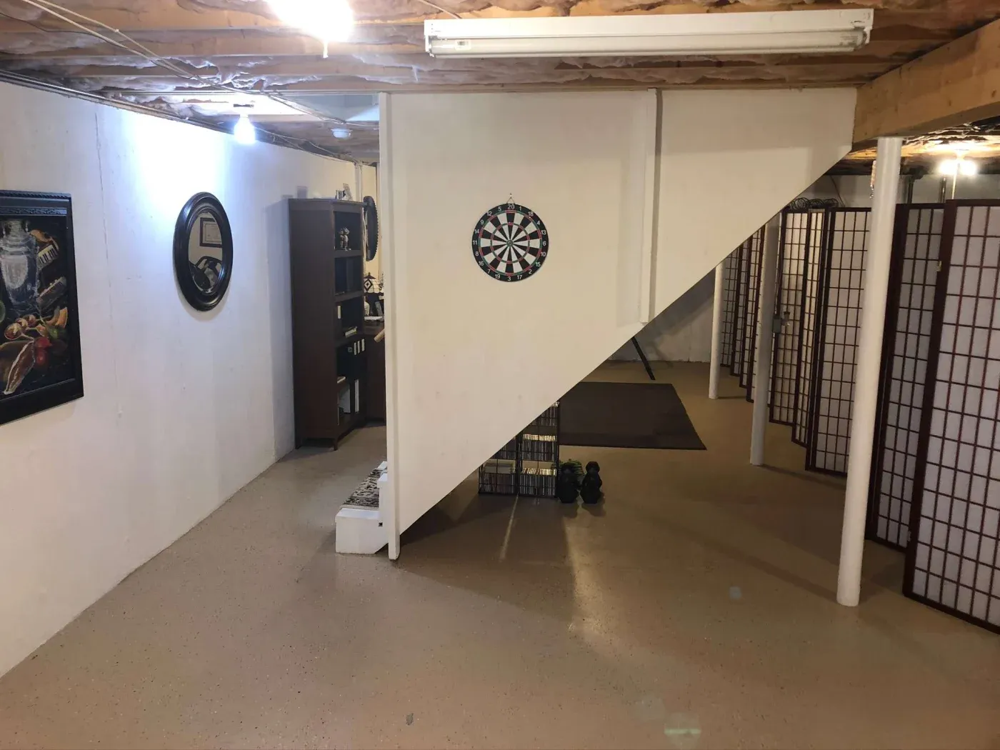 A basement setup with a dartboard on a white wall under a staircase. Cozy atmosphere with a bookcase, framed artwork, and room dividers.
