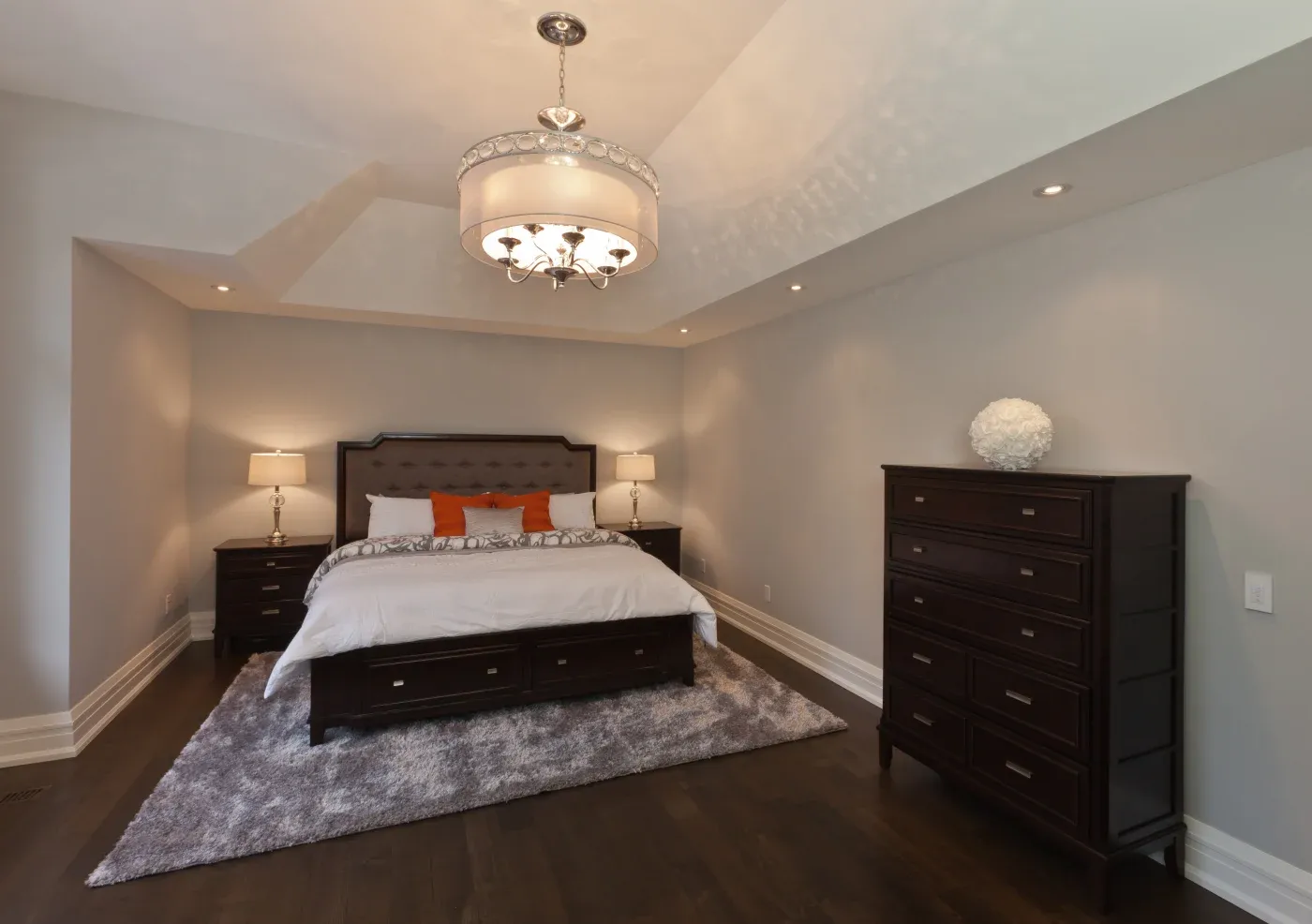 Spacious bedroom with a large bed featuring white and orange pillows. Dark wood furniture, soft lighting, gray rug, and an elegant chandelier create a cozy atmosphere.