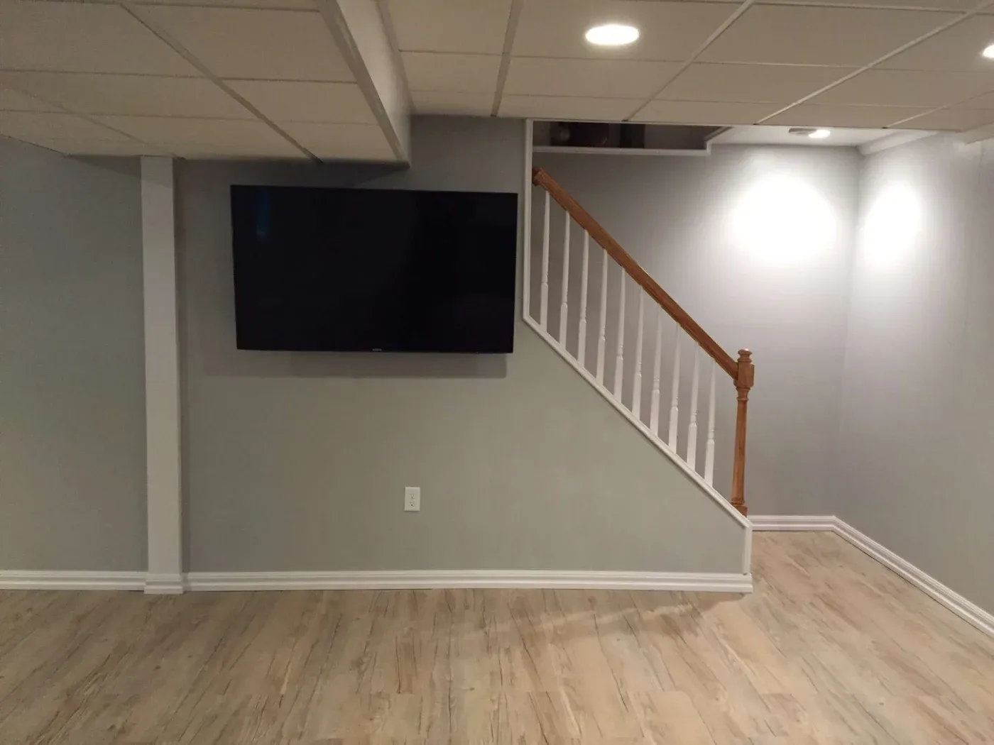 A modern basement with light wood flooring and gray walls features a large flat-screen TV on the wall. A staircase with wooden handrails is on the right.