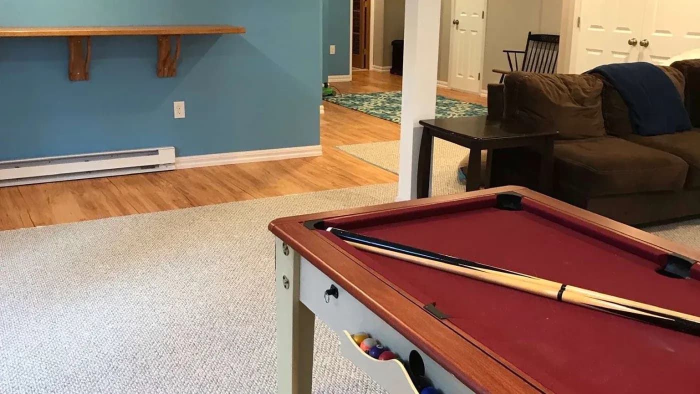 A cozy game room featuring a red pool table with a cue stick, beige carpet, blue walls, and a brown sofa. The ambiance is inviting and leisure-focused.
