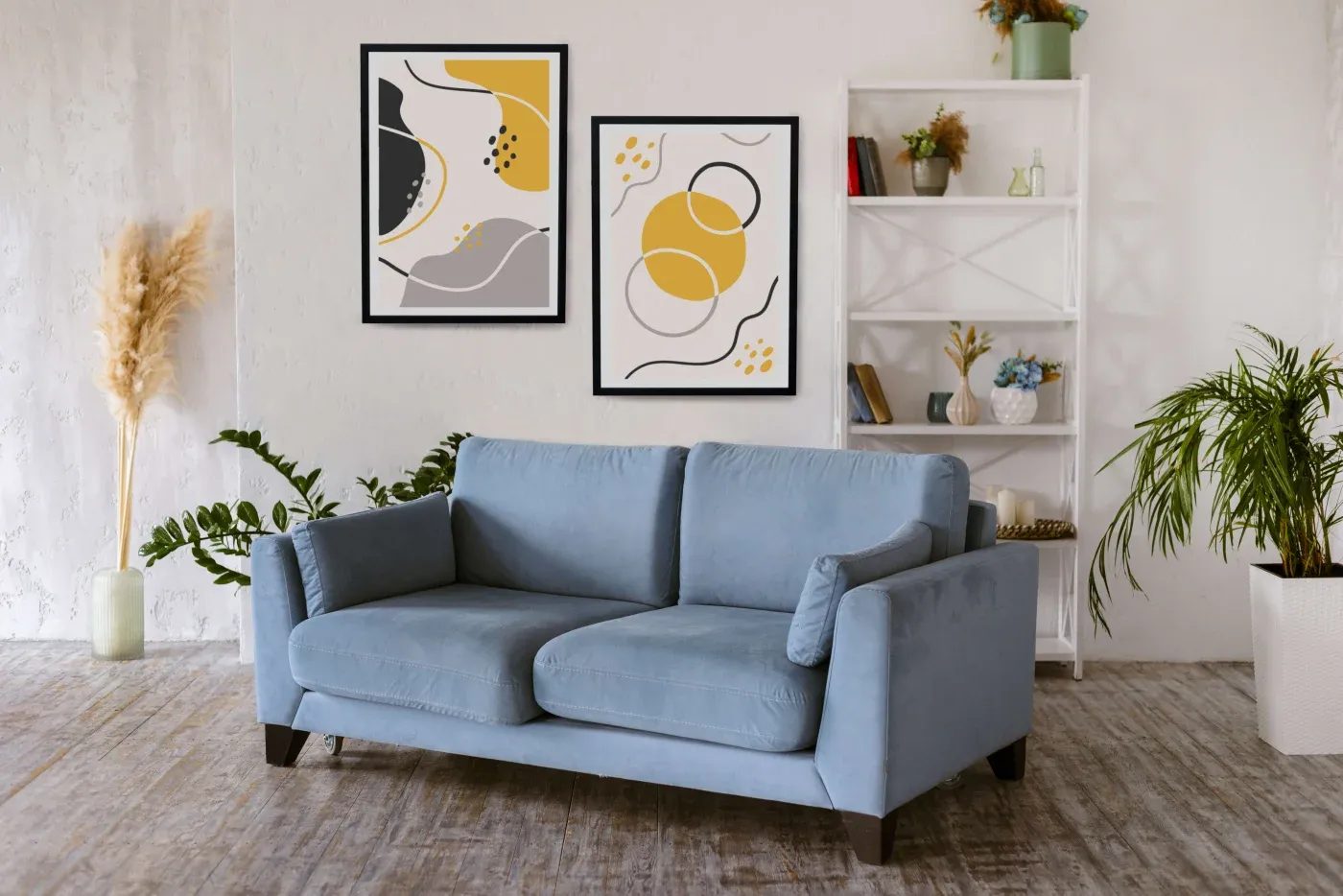 A cozy living room features a light blue sofa against a white wall, adorned with two abstract paintings in black, yellow, and gray. Nearby, a white shelf holds books and decor, with plants adding a touch of greenery.